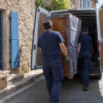 Deux intervenants chargent une armoire protégée par des couvertures dans un utilitaire devant une maison de village de l’Hérault